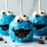 Blue Cookie Monster-themed candy apples with edible candy eyes, a wide cookie-filled mouth, and a glossy candy coating, placed on a marble surface with scattered Oreo crumbs.