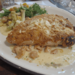 A plate of Longhorn Steakhouse Parmesan Crusted Chicken served with creamy garlic sauce and a side of mixed vegetables. The chicken is golden brown, crispy, and topped with melted parmesan and crunchy breadcrumbs.