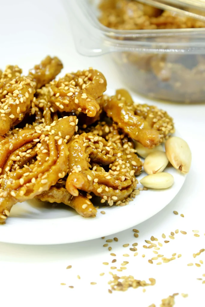 A close-up of Moroccan Chebakia, a traditional honey-glazed pastry sprinkled with sesame seeds, served on a white plate with peanuts on the side.